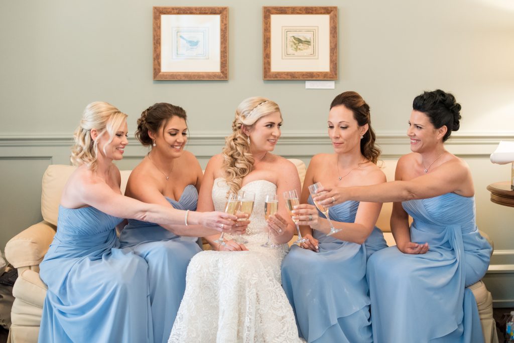 bride sits with her bridesmaids wearing powder blue dresses holding champagne glasses together while on couch
